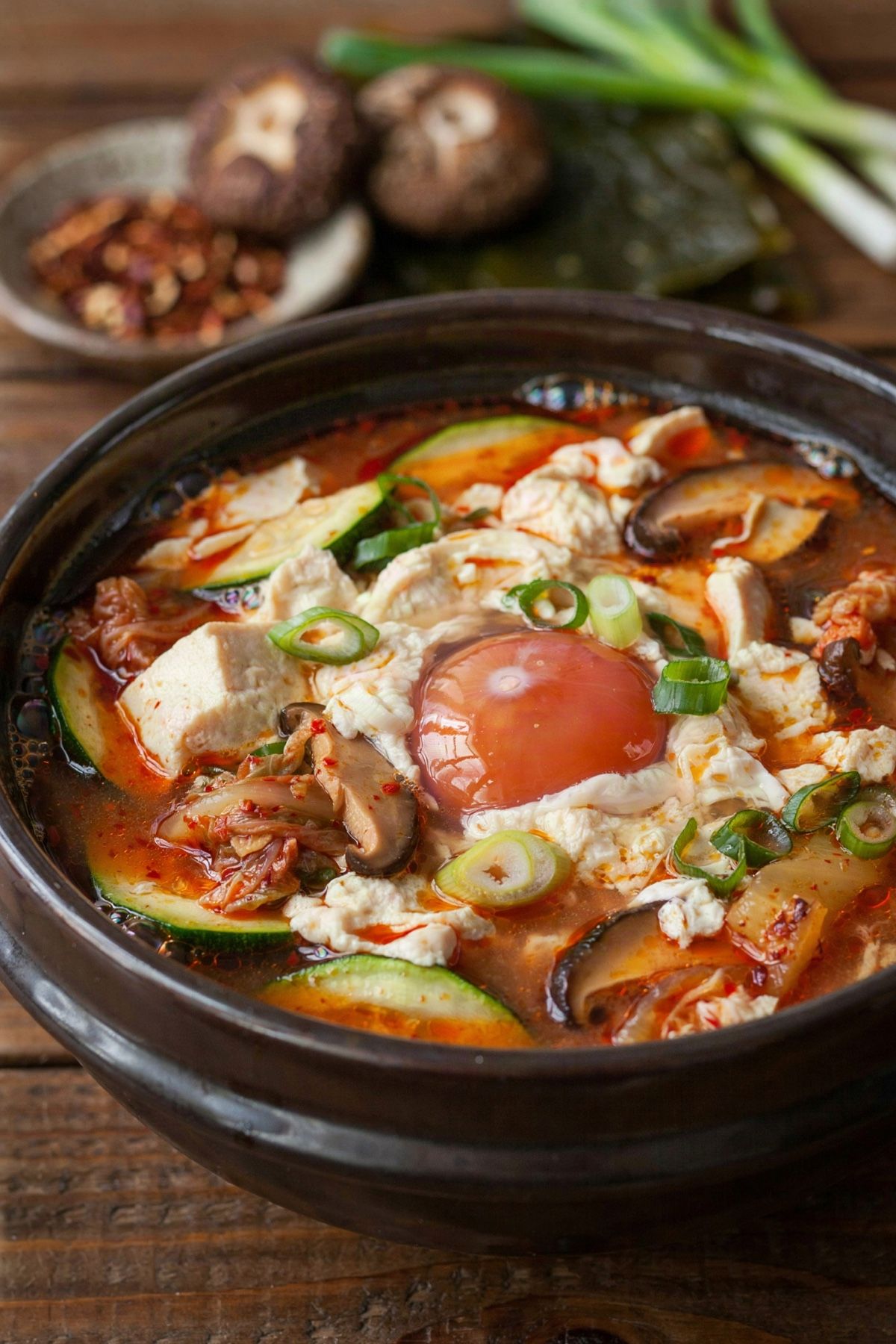 Vegetarian sundubu jjigae in a dark earthenware bowl, soft curds of silky tofu in a vivid red chili broth with zucchini, kimchi and shiitake, one soft-yolked egg in the center and sliced scallions on top.