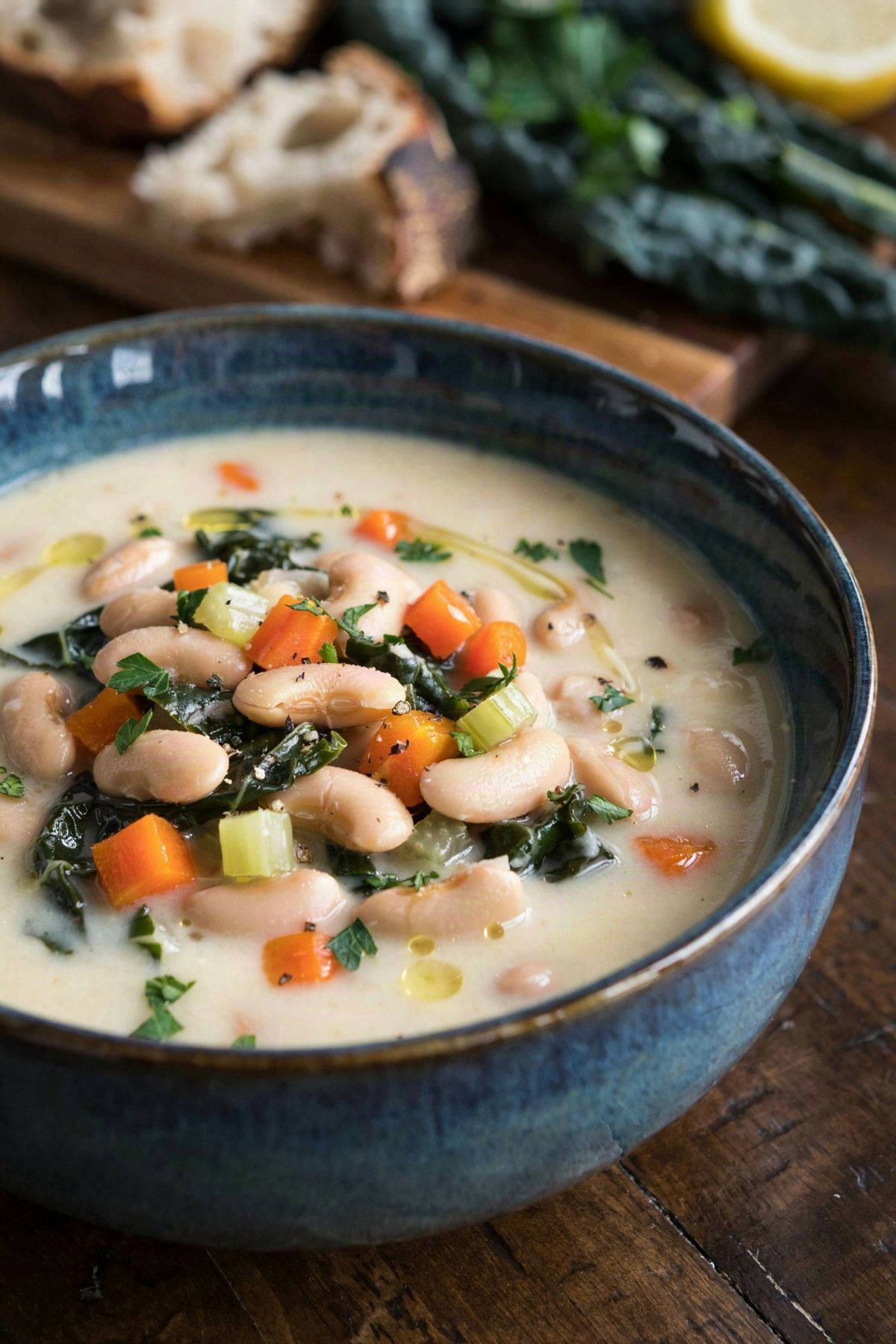 Vegan white bean soup in a blue glazed bowl, thick and creamy pale ivory from the crushed beans, with whole cannellini, diced carrot and ribbons of dark green kale, finished with a drizzle of olive oil and chopped parsley.