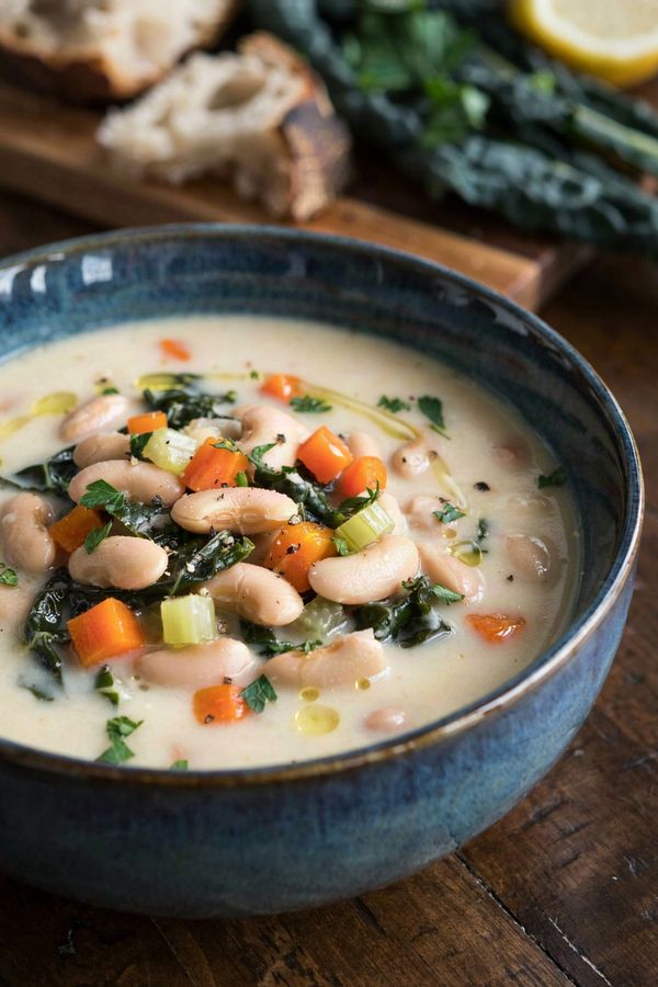 Vegan white bean soup in a blue glazed bowl, thick and creamy pale ivory from the crushed beans, with whole cannellini, diced carrot and ribbons of dark green kale, finished with a drizzle of olive oil and chopped parsley.