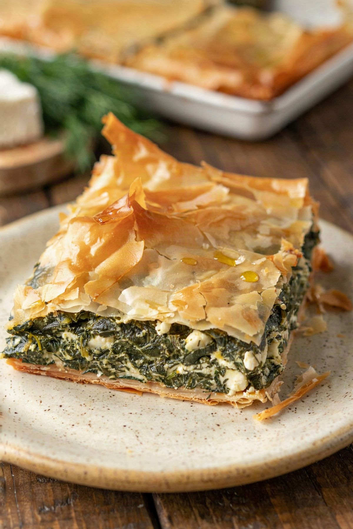 A square of Greek spanakopita on a cream plate, flaky golden phyllo over a thick green spinach and feta filling on a crisp golden base, with the rest of the pie behind.