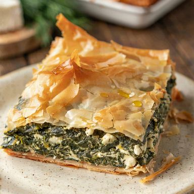 A square of Greek spanakopita on a cream plate, flaky golden phyllo over a thick green spinach and feta filling on a crisp golden base, with the rest of the pie behind.