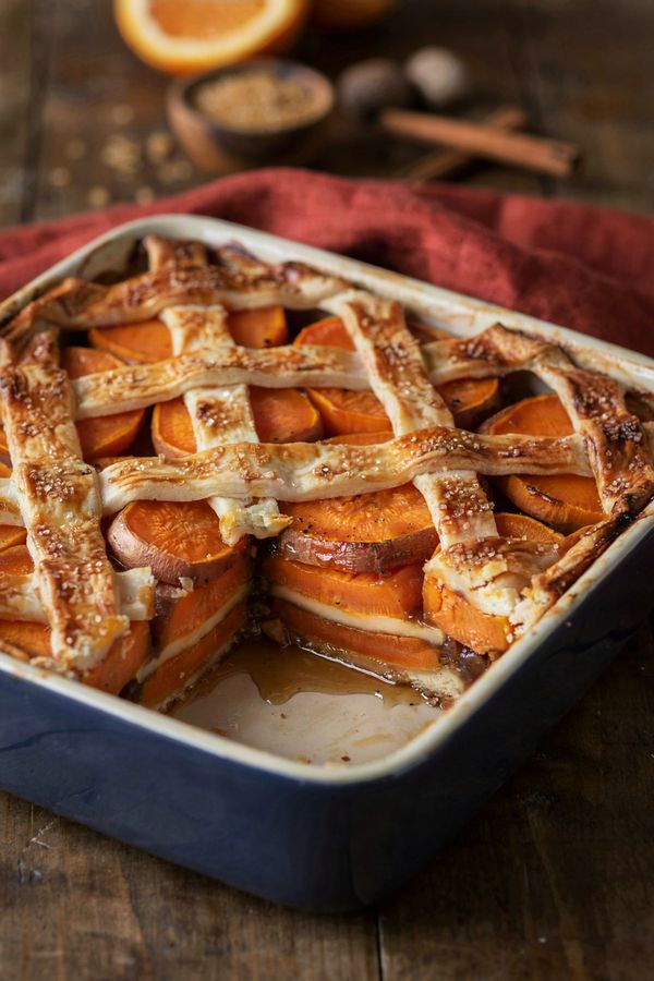 Sweet potato cobbler in a navy baking dish, a golden pastry lattice over sliced sweet potatoes in dark amber syrup, with one corner spooned out to show the pastry layer baked through the middle