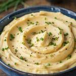 Vegan mashed potatoes swirled into soft ridges in a deep blue stoneware bowl, golden and smooth, scattered with snipped chives and cracked black pepper.