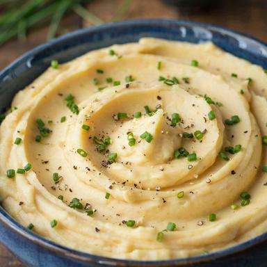 Vegan mashed potatoes swirled into soft ridges in a deep blue stoneware bowl, golden and smooth, scattered with snipped chives and cracked black pepper.