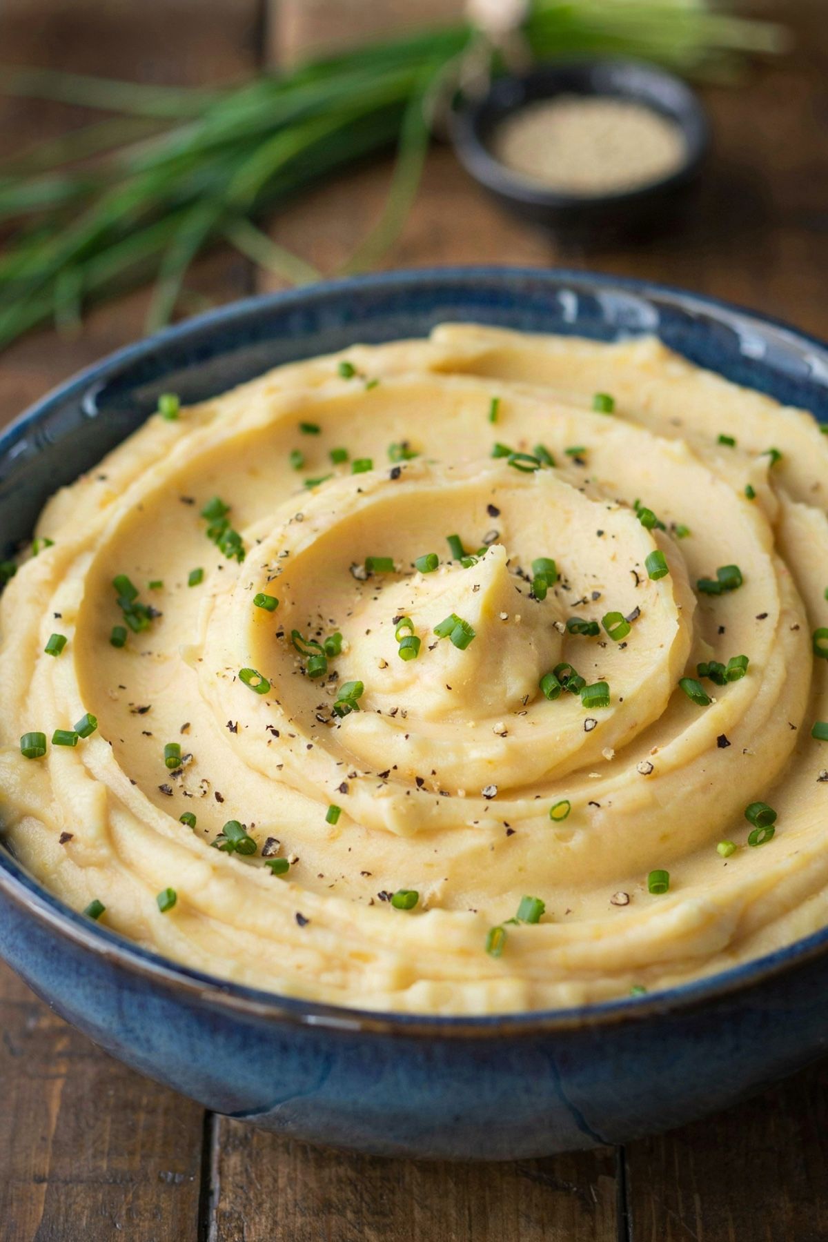 Vegan mashed potatoes swirled into soft ridges in a deep blue stoneware bowl, golden and smooth, scattered with snipped chives and cracked black pepper.