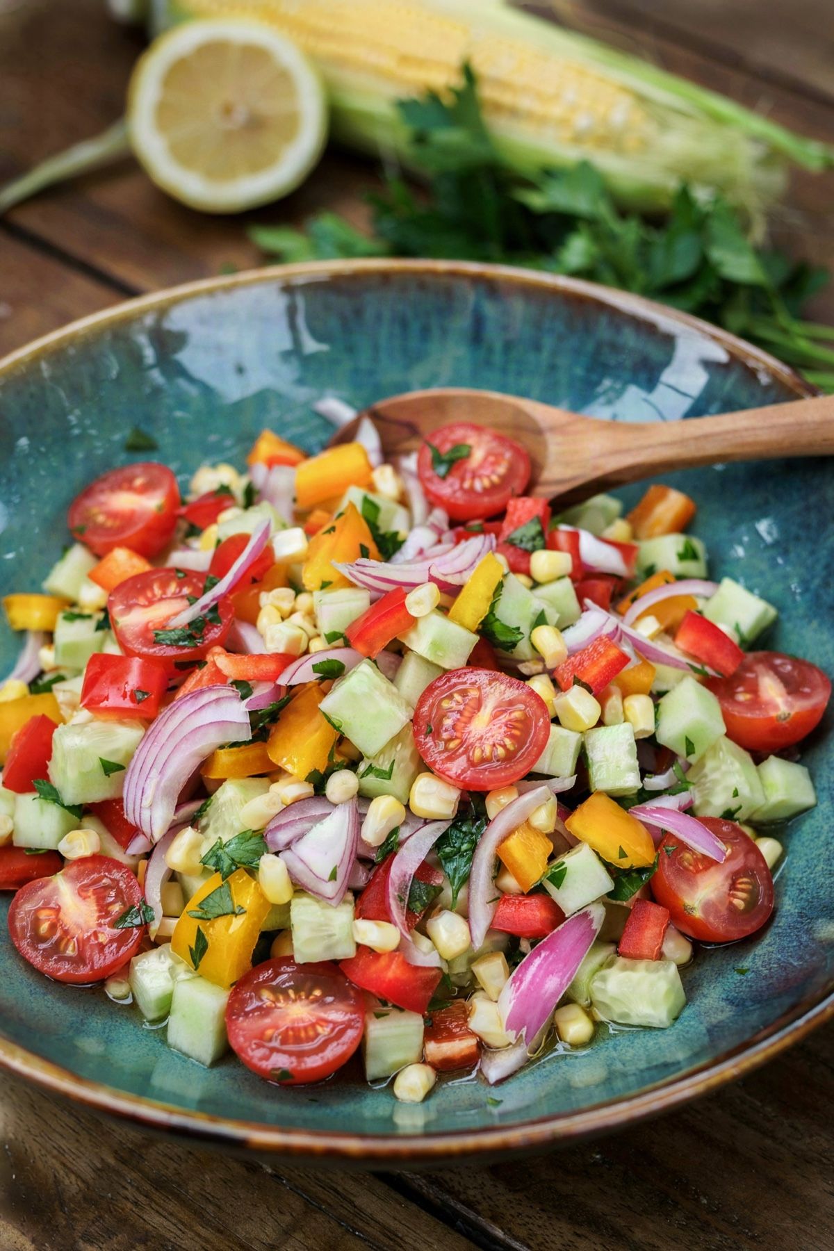 Chopped veggie salad without lettuce in a teal bowl: halved cherry tomatoes, diced cucumber, red, yellow and orange bell pepper, sweetcorn, thin pink red onion and parsley glossy with lemon and olive oil dressing.