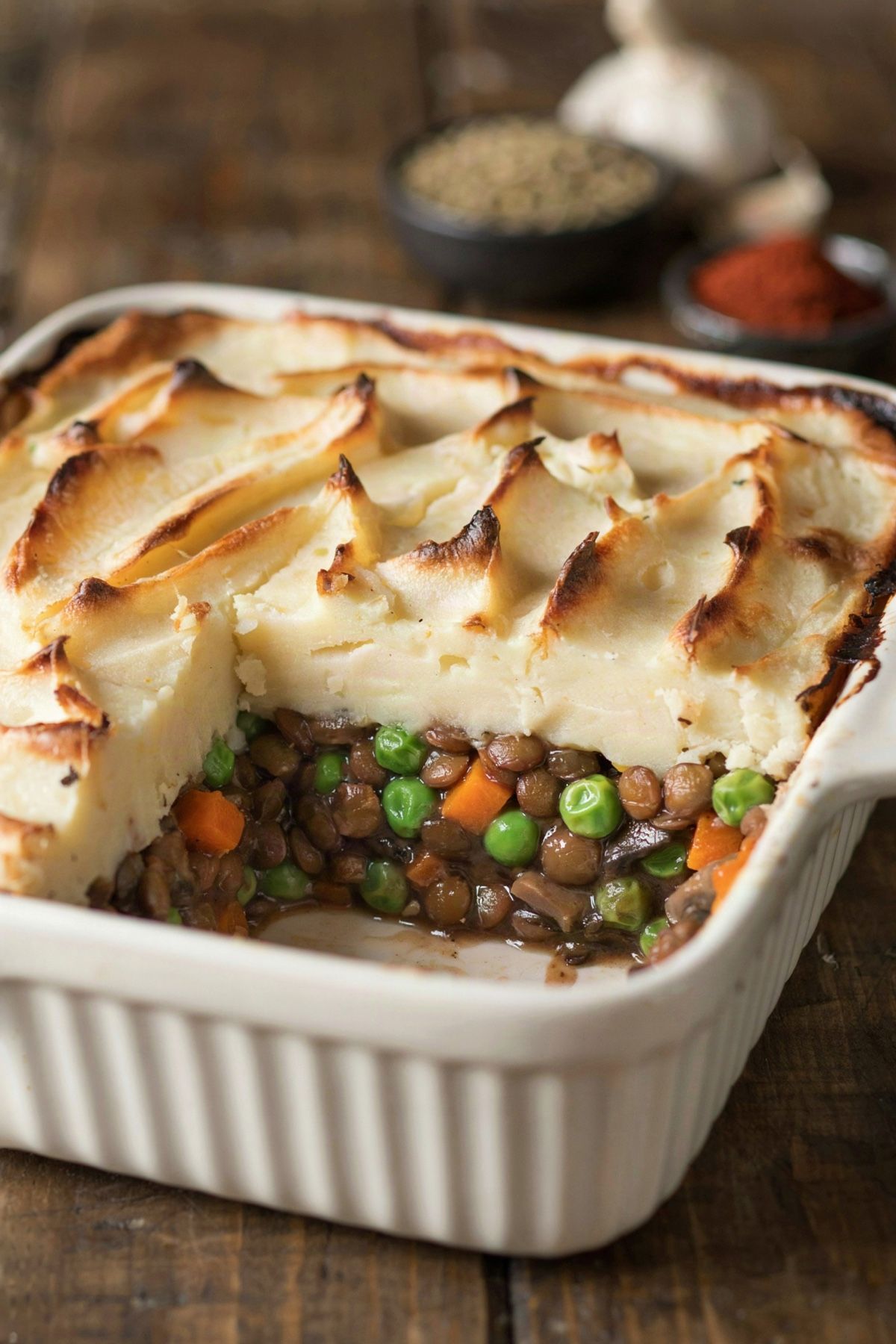 Lentil cottage pie in a white ceramic dish, a square portion lifted out to show brown lentils, mushrooms, peas and carrot in gravy under a golden forked mash lid