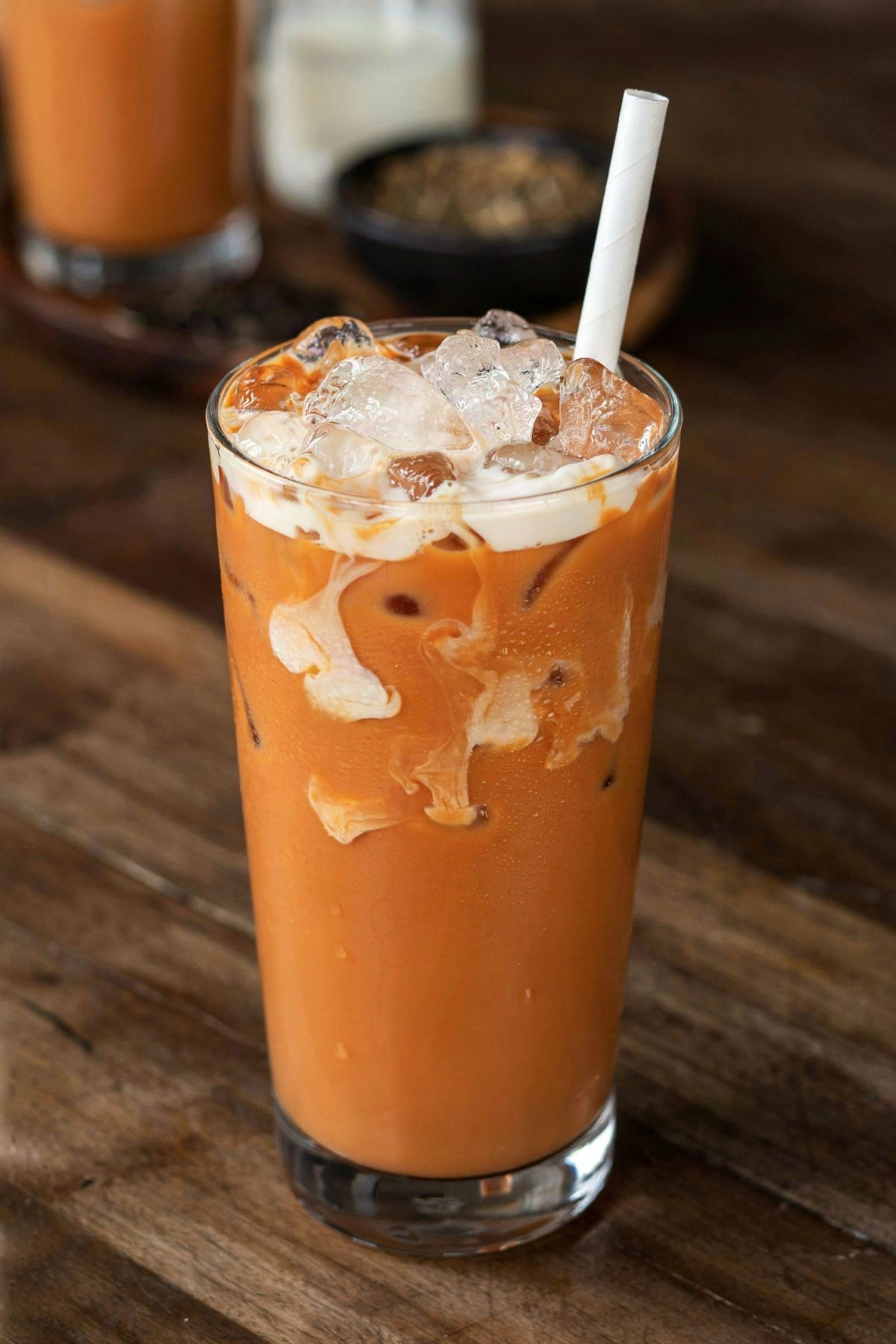 A tall glass of Thai iced tea, bright orange milk tea over ice with evaporated milk drifting down from the top in white clouds and a paper straw.
