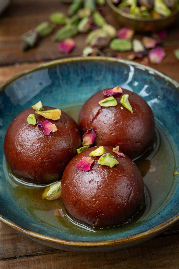 Three gulab jamun in a deep teal bowl, round reddish brown milk dumplings half submerged in thin golden cardamom and rose syrup, scattered with slivered pistachios and dried rose petals.