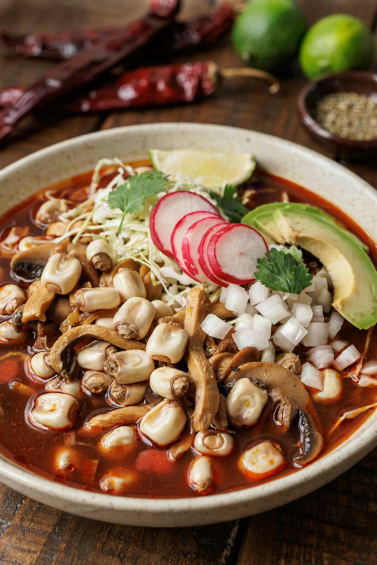 Vegan pozole rojo in a speckled stoneware bowl, hominy kernels and torn oyster mushrooms in a deep red chile broth, topped with shredded cabbage, radish, white onion, cilantro and avocado.