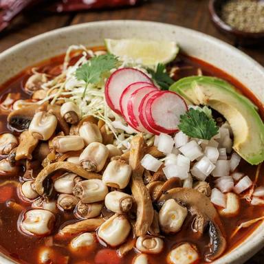 Vegan pozole rojo in a speckled stoneware bowl, hominy kernels and torn oyster mushrooms in a deep red chile broth, topped with shredded cabbage, radish, white onion, cilantro and avocado.