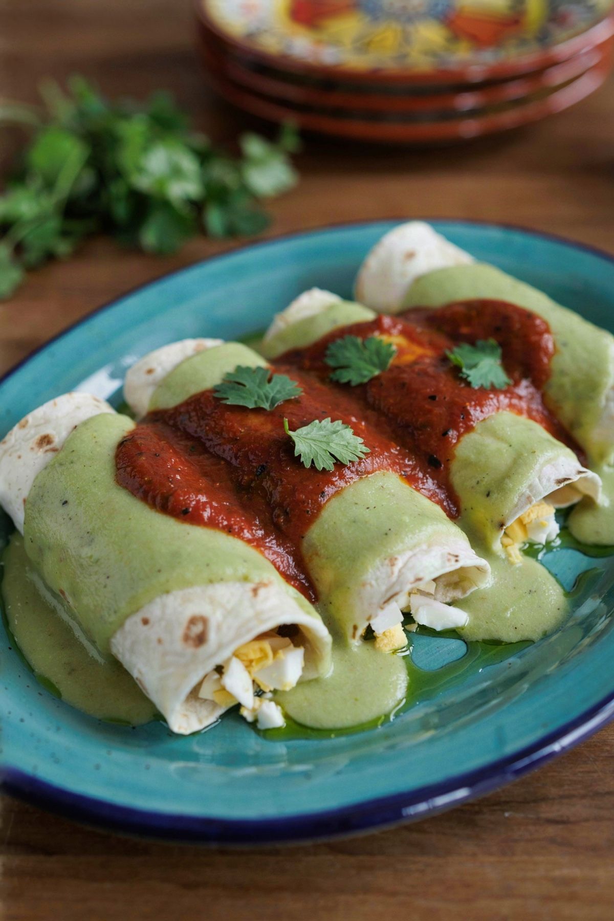 Three papadzules on a turquoise plate, corn tortillas rolled around chopped egg under a pale green pumpkin seed sauce with brick red chiltomate and cilantro.