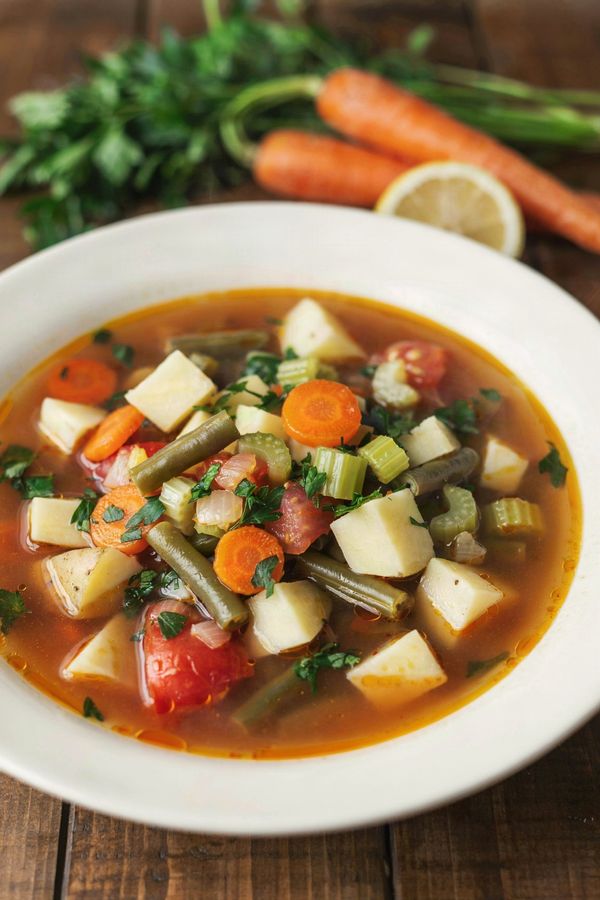 A white bowl of Instant Pot vegetable soup in a red-orange broth, with cubes of potato, carrot coins, sliced celery, green beans and soft tomato, scattered with fresh parsley.