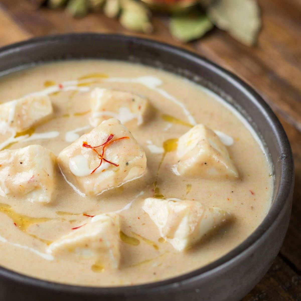 Shahi paneer in a dark stoneware bowl, soft paneer cubes in a pale ivory Mughlai cream gravy with a swirl of cream and a few saffron threads on top.