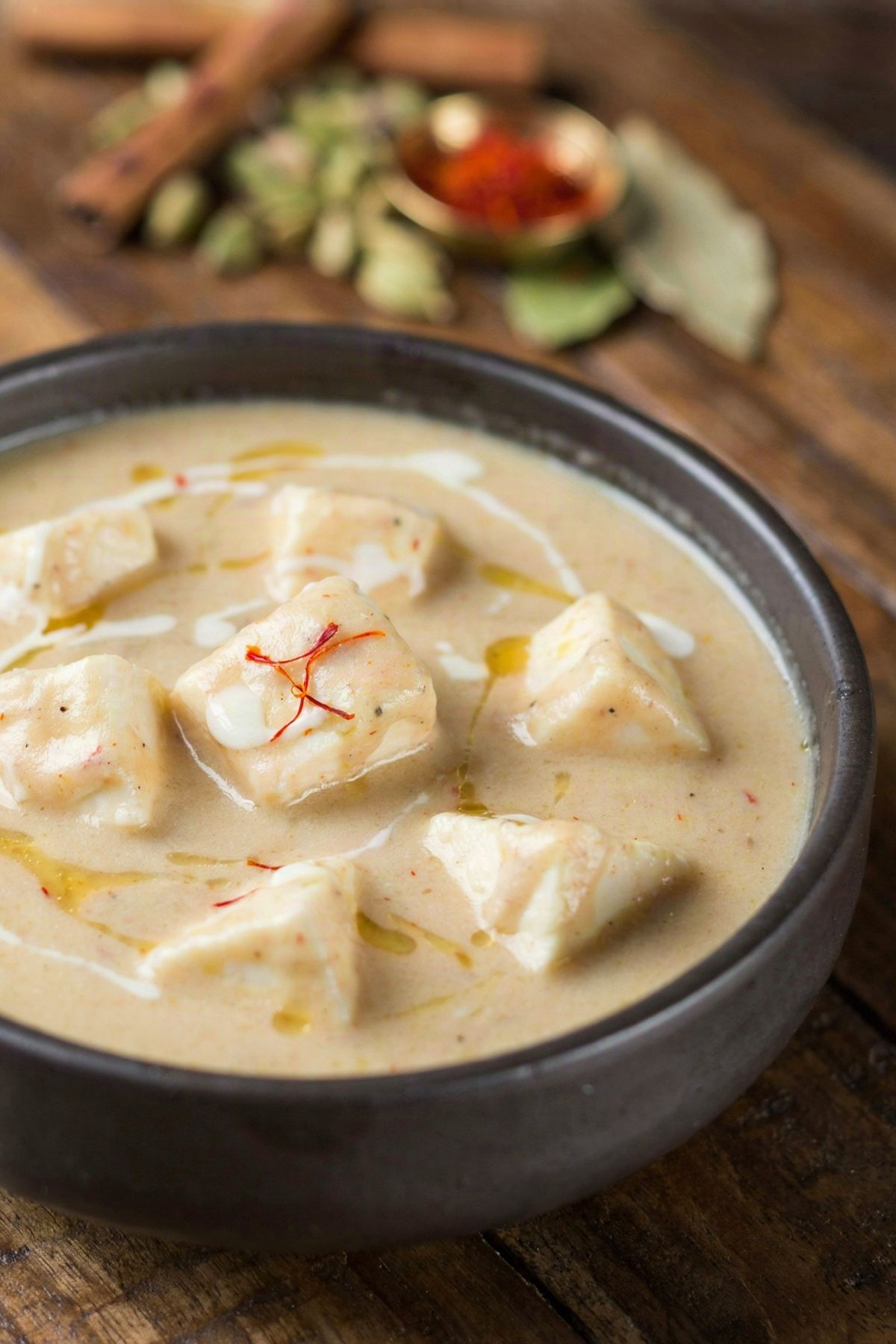 Shahi paneer in a dark stoneware bowl, soft paneer cubes in a pale ivory Mughlai cream gravy with a swirl of cream and a few saffron threads on top.
