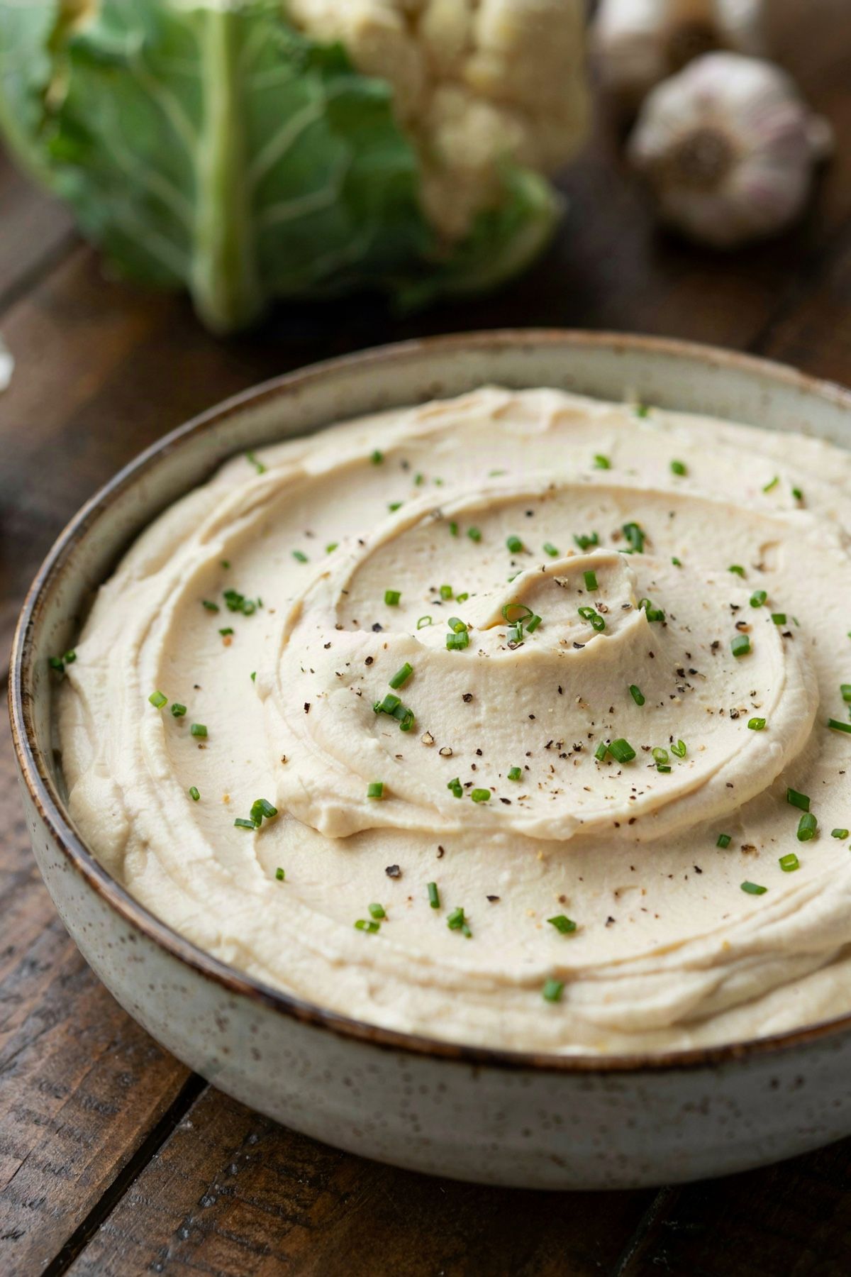 Creamy mashed cauliflower in a speckled stoneware bowl, swirled thick and pale and scattered with snipped chives and coarse black pepper, a whole cauliflower and garlic bulbs on the table behind.