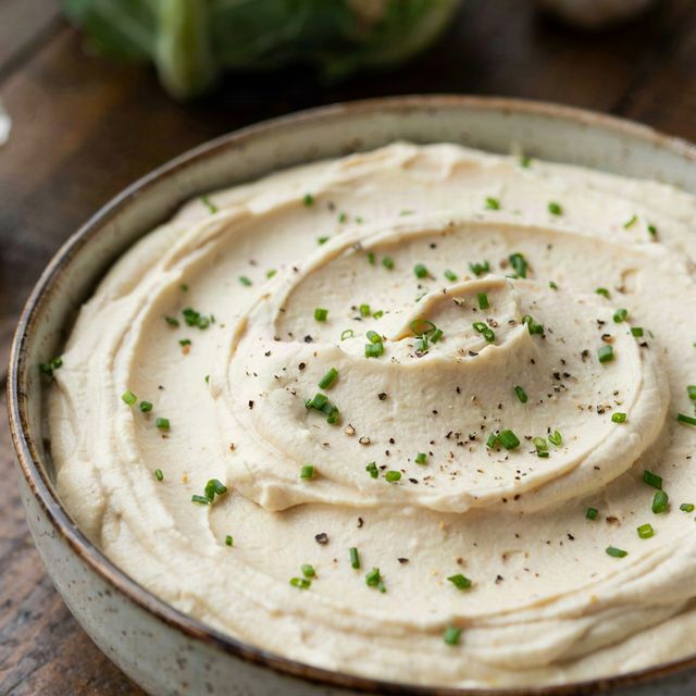 Creamy mashed cauliflower in a speckled stoneware bowl, swirled thick and pale and scattered with snipped chives and coarse black pepper, a whole cauliflower and garlic bulbs on the table behind.