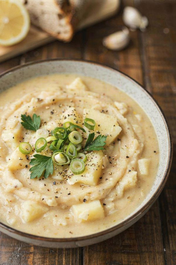 Thick vegan potato soup in a speckled stoneware bowl, mashed rather than blended so soft chunks of potato show through, topped with sliced green onion, parsley and cracked black pepper.