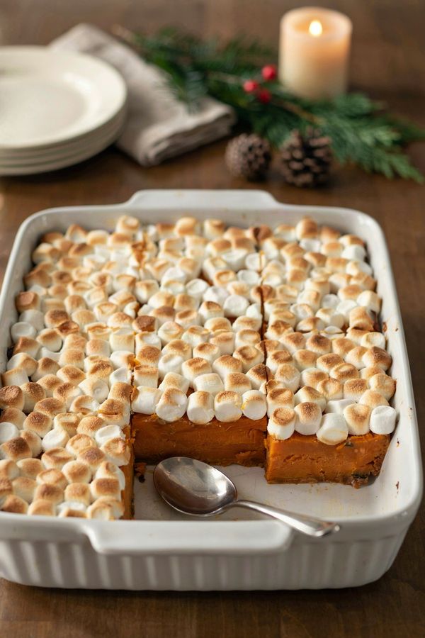 Sweet potato casserole in a white baking dish on a candlelit holiday table, deep orange baked sweet potato under a layer of golden toasted mini marshmallows, with one serving scooped out to show the set layers