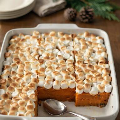 Sweet potato casserole in a white baking dish on a candlelit holiday table, deep orange baked sweet potato under a layer of golden toasted mini marshmallows, with one serving scooped out to show the set layers
