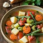 A bowl of homemade vegetable soup in a tomato broth with potato, carrot, celery, bright green beans and tomato, scattered with parsley and served with crusty bread.