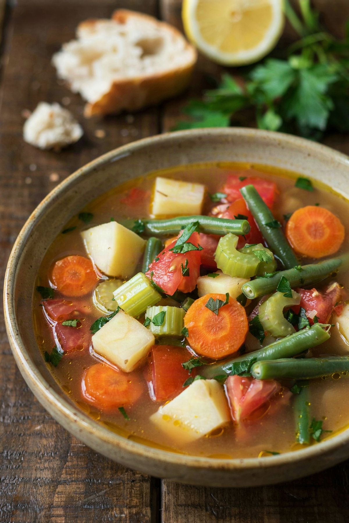 A bowl of homemade vegetable soup in a tomato broth with potato, carrot, celery, bright green beans and tomato, scattered with parsley and served with crusty bread.