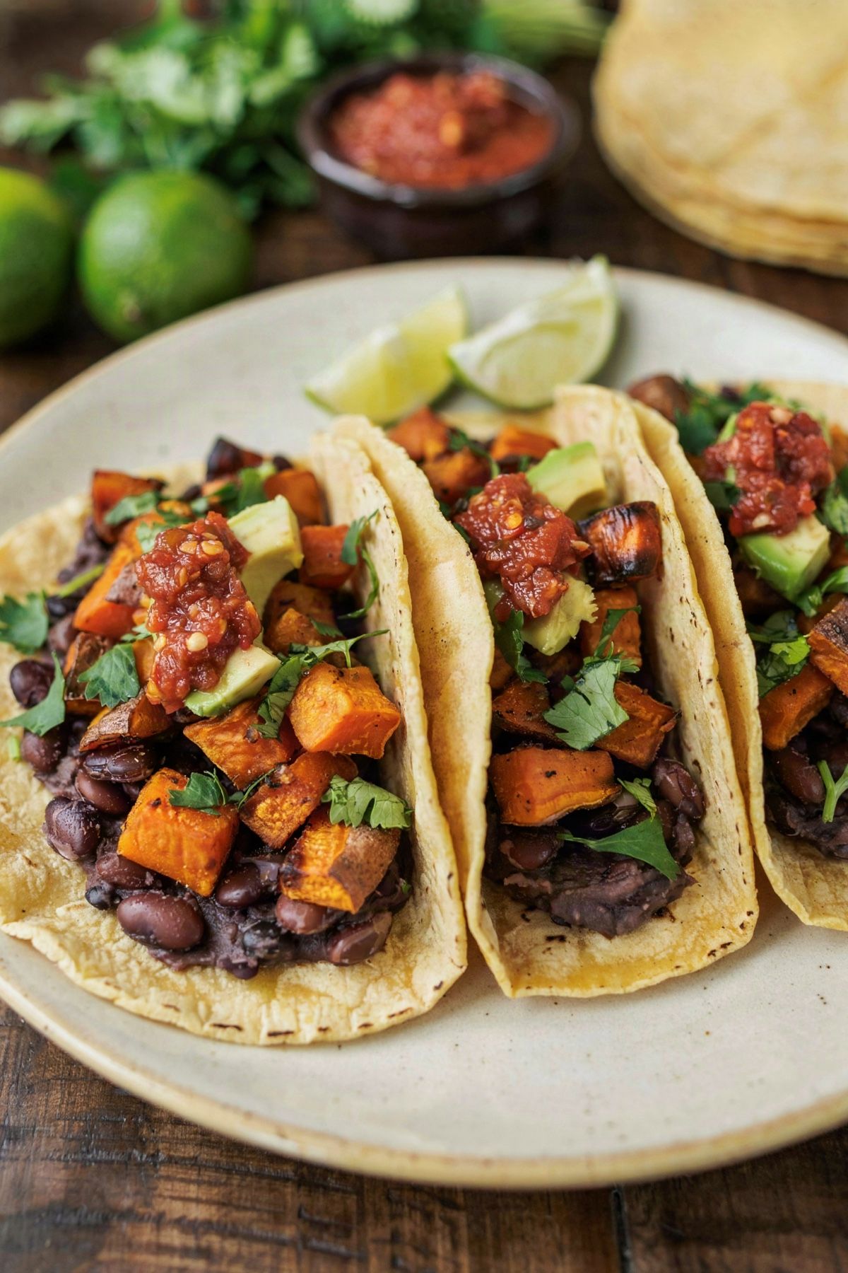 Three sweet potato and black bean tacos on a cream plate, soft yellow corn tortillas spread with dark mashed black beans and piled with caramelized roasted sweet potato, brick red salsa, avocado and cilantro.