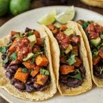 Three sweet potato and black bean tacos on a cream plate, soft yellow corn tortillas spread with dark mashed black beans and piled with caramelized roasted sweet potato, brick red salsa, avocado and cilantro.