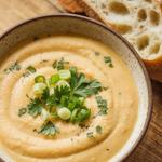 A bowl of creamy vegan potato soup garnished with green onions and parsley, with a slice of bread on the side.