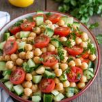 A vibrant vegan chickpea salad with cucumbers, tomatoes, and parsley on a wooden table with lemon.