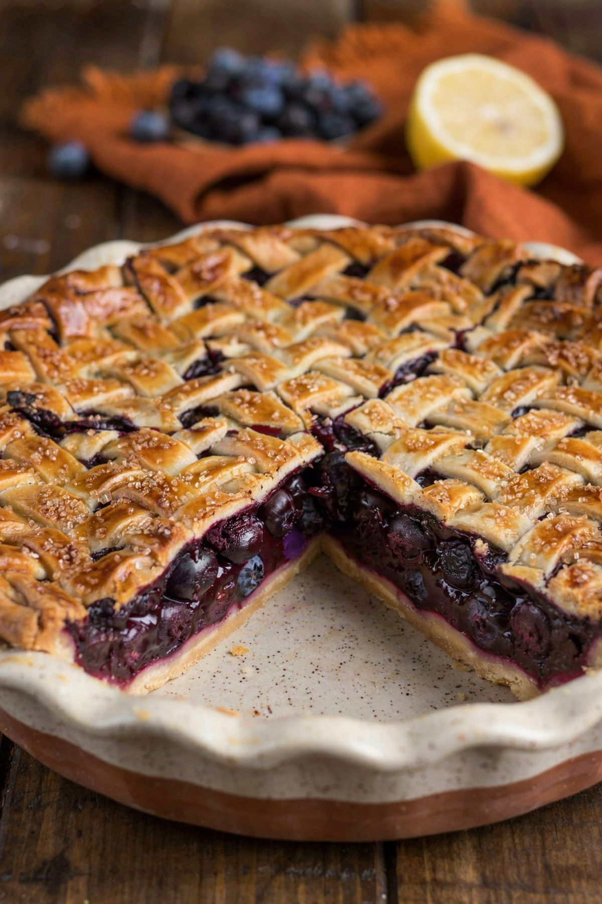 A vegan blueberry pie in a white ceramic dish with one slice cut away, the dark purple filling set firm under a golden lattice crust