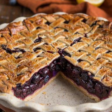 A vegan blueberry pie in a white ceramic dish with one slice cut away, the dark purple filling set firm under a golden lattice crust