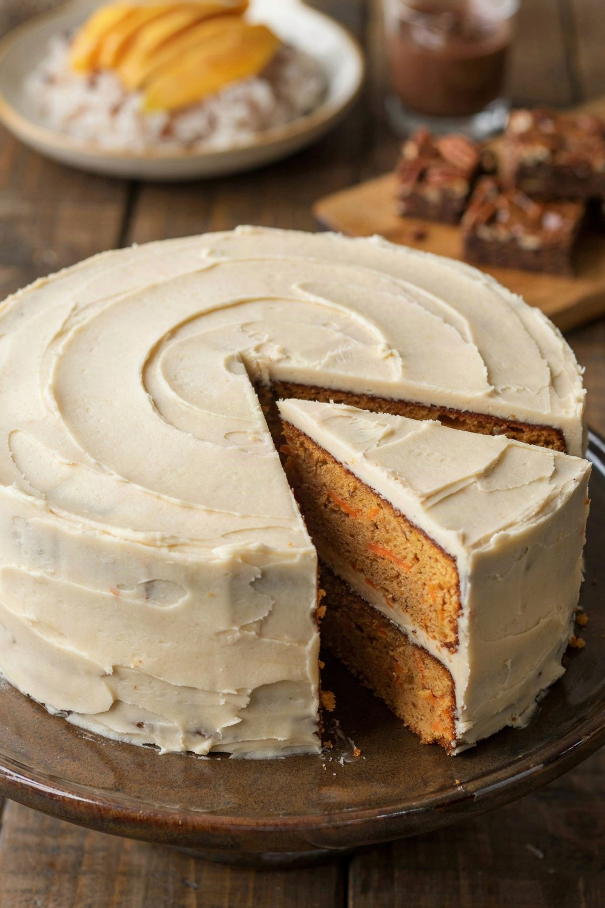 A two-layer vegan carrot cake under off-white frosting on a dark stand, one slice cut to show the carrot-flecked crumb, with mango sticky rice, chocolate mousse and pecan bars out of focus on the table behind.