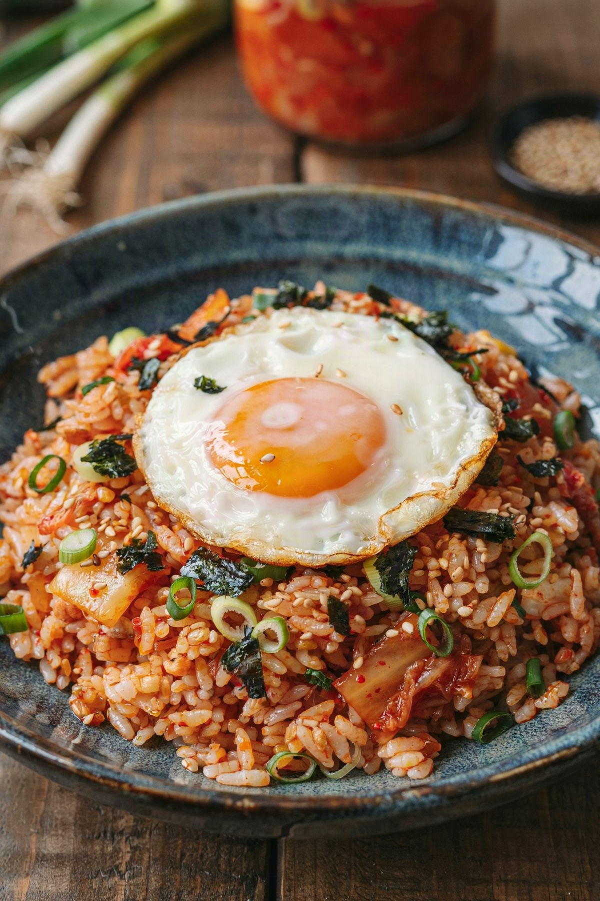 Kimchi fried rice in a dark blue-gray ceramic bowl, orange-red rice flecked with fried kimchi under a sunny side up egg with a runny yolk, scattered with scallion greens, sesame seeds and crumbled roasted seaweed.