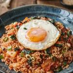 Kimchi fried rice in a dark blue-gray ceramic bowl, orange-red rice flecked with fried kimchi under a sunny side up egg with a runny yolk, scattered with scallion greens, sesame seeds and crumbled roasted seaweed.