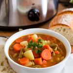 A bowl of vegetable crock pot soup with carrots, potatoes, green beans, garnished with parsley, alongside crusty bread.