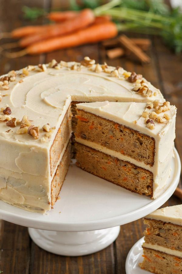 A two-layer vegan carrot cake on a white cake stand, covered in ivory cream cheese frosting with chopped walnuts around the top edge and one wedge cut to show the tan crumb flecked with carrot.