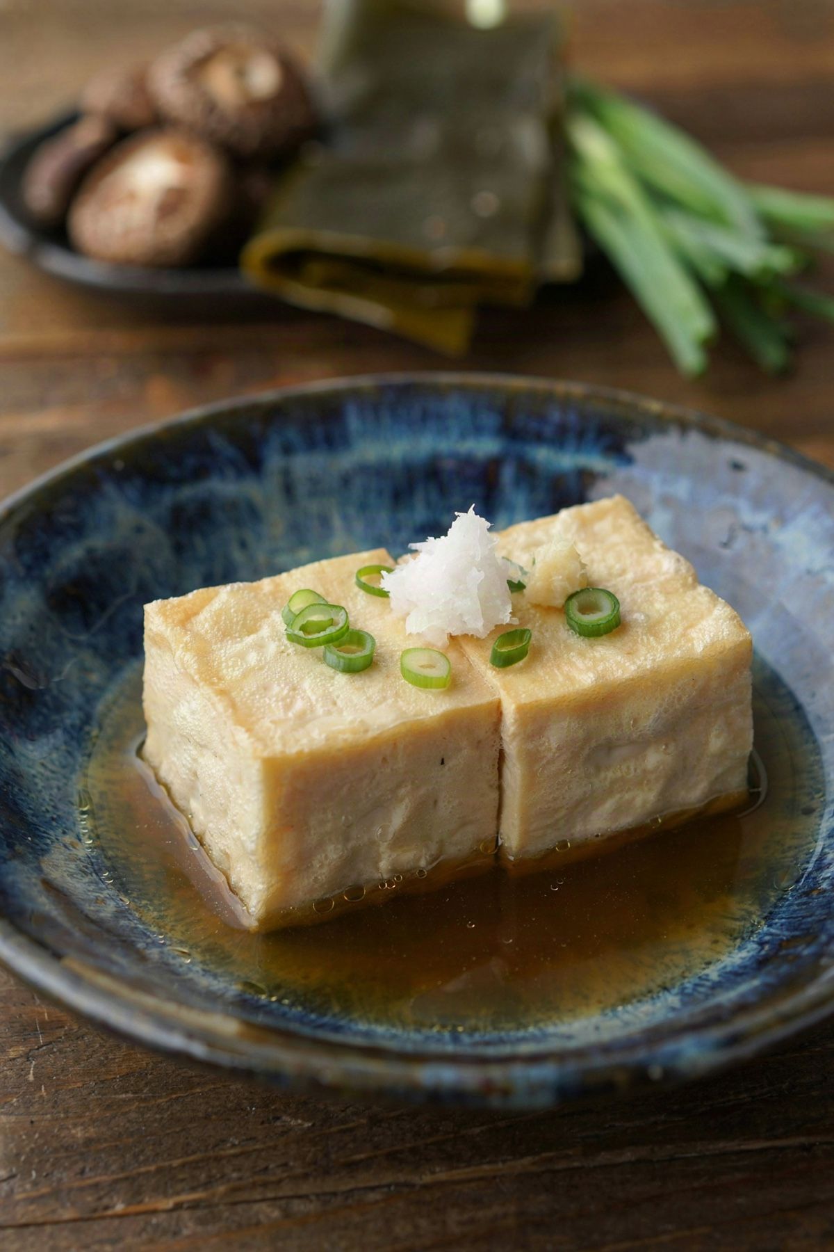 Agedashi tofu in a dark blue bowl, two cubes of tofu in a thin pale gold starch shell standing in clear amber dashi broth, topped with grated daikon, ginger and sliced scallion.