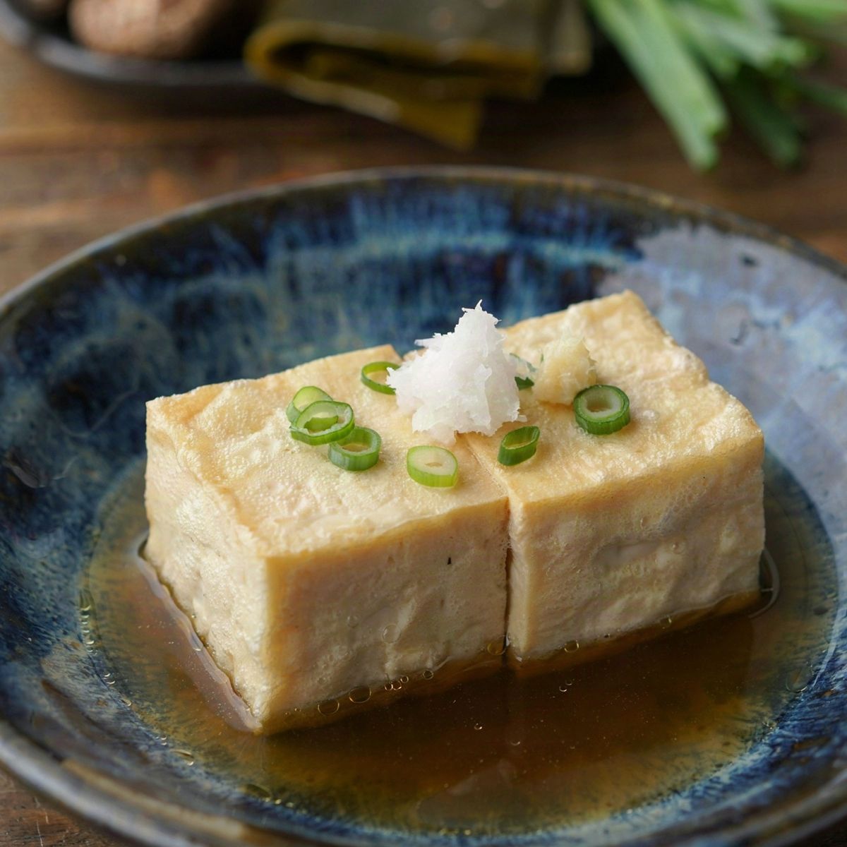 Agedashi tofu in a dark blue bowl, two cubes of tofu in a thin pale gold starch shell standing in clear amber dashi broth, topped with grated daikon, ginger and sliced scallion.