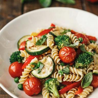 Vegan primavera pasta in a white bowl: fusilli with red bell pepper, zucchini, blistered cherry tomatoes, broccoli and peas, finished with torn basil