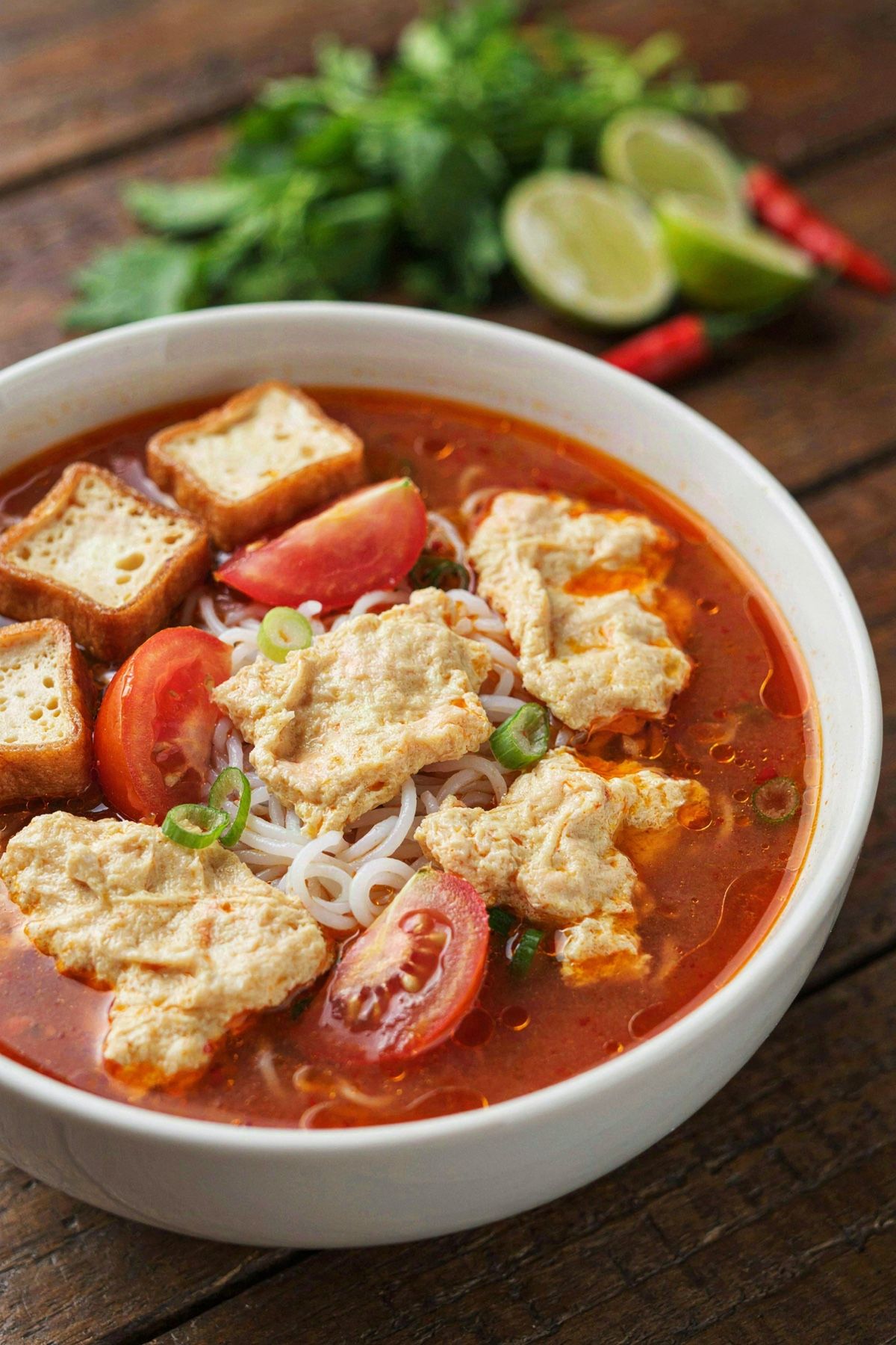 A bowl of vegetarian bun rieu, orange-red tomato broth with ragged golden curds, rice vermicelli, tomato wedges and fried tofu puffs, with fresh herbs and lime alongside.