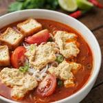 A bowl of vegetarian bun rieu, orange-red tomato broth with ragged golden curds, rice vermicelli, tomato wedges and fried tofu puffs, with fresh herbs and lime alongside.