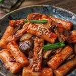 Vegetarian tteokbokki in a shallow dark bowl, cylindrical Korean rice cakes coated in a thick glossy deep red gochujang sauce with fried tofu puff strips, sliced shiitake, onion, scallion and toasted sesame seeds.