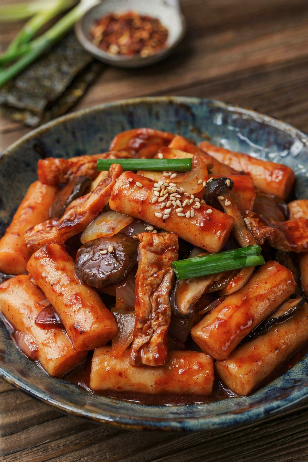 Vegetarian tteokbokki in a shallow dark bowl, cylindrical Korean rice cakes coated in a thick glossy deep red gochujang sauce with fried tofu puff strips, sliced shiitake, onion, scallion and toasted sesame seeds.