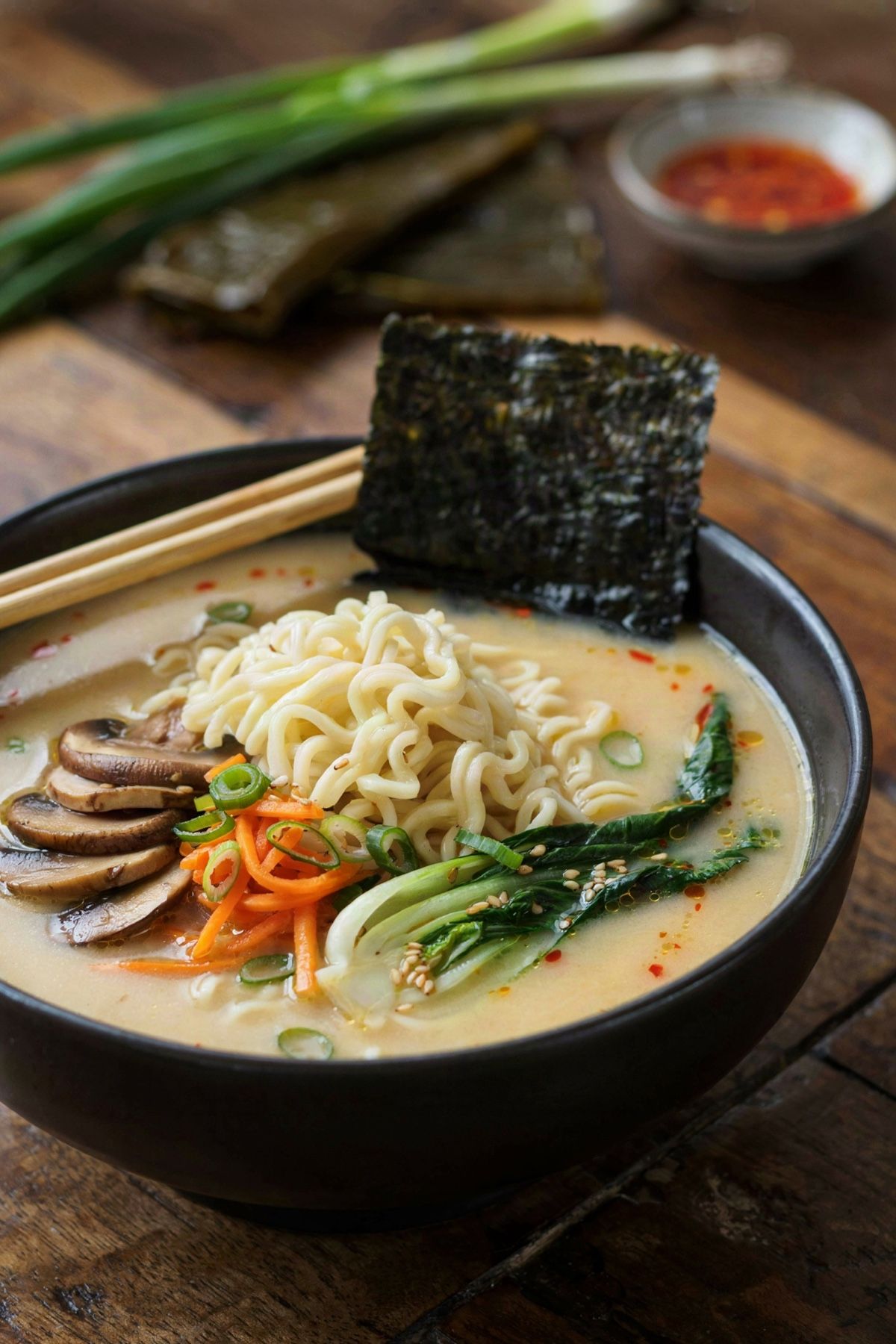 Vegan ramen in a black bowl, wavy noodles with shiitake, carrot matchsticks and baby bok choy in an opaque miso broth, with a sheet of nori at the rim.