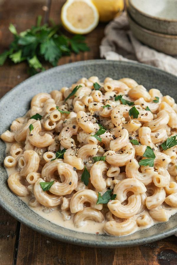 Creamy vegan mac and cheese in a speckled stoneware bowl, ridged elbow macaroni coated in a pale beige cashew and nutritional yeast sauce, scattered with chopped parsley and cracked black pepper.