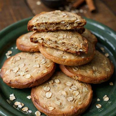 Vegan oatmeal cookies stacked on a green stoneware plate, thick golden discs packed with rolled oats, one broken open to show the chewy inside