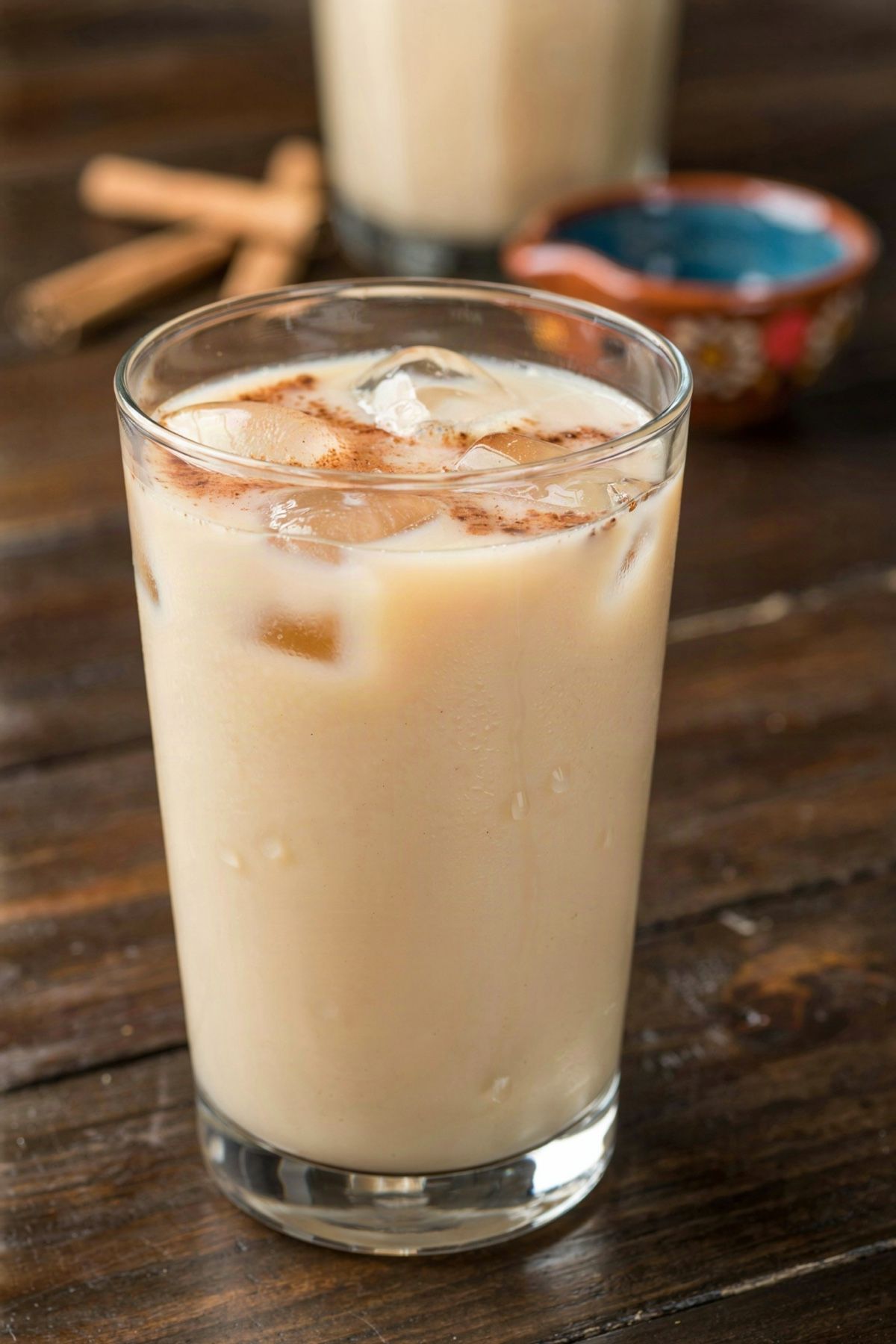 A tall glass of Mexican horchata over ice on a dark wooden table, the pale ivory rice drink dusted with ground cinnamon, with a pitcher of it and loose canela sticks behind.