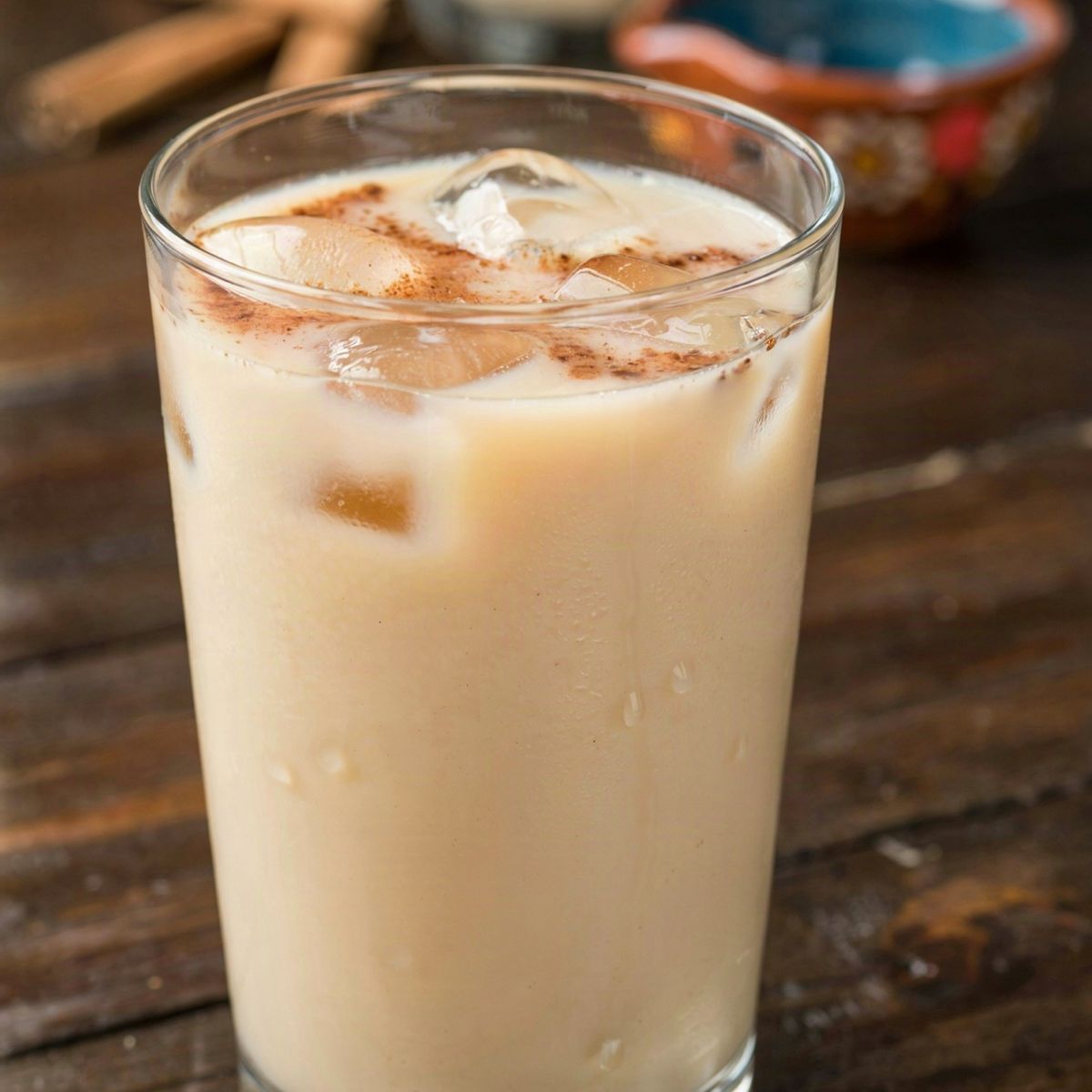 A tall glass of Mexican horchata over ice on a dark wooden table, the pale ivory rice drink dusted with ground cinnamon, with a pitcher of it and loose canela sticks behind.