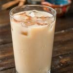 A tall glass of Mexican horchata over ice on a dark wooden table, the pale ivory rice drink dusted with ground cinnamon, with a pitcher of it and loose canela sticks behind.